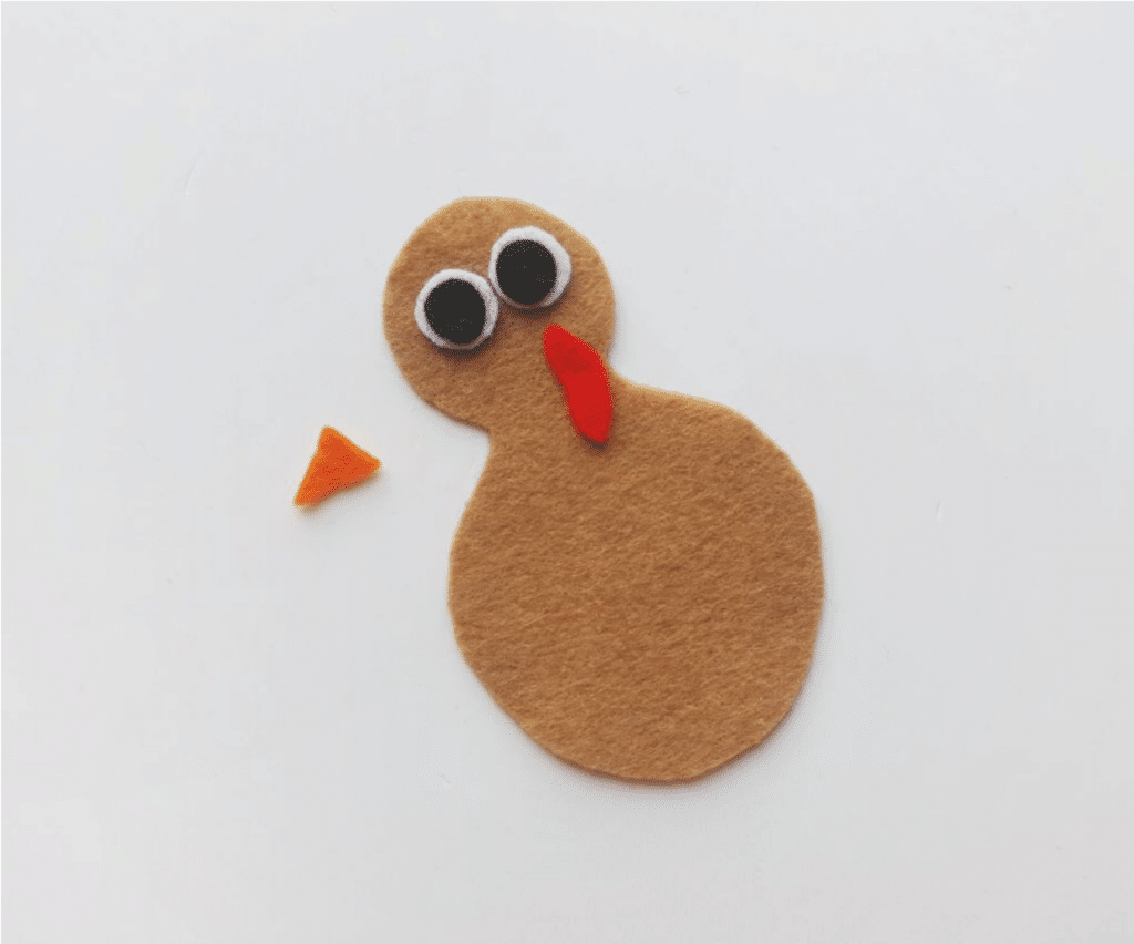 A felt cutout of a brown turkey with black and white eyes and a red wattle, perfect for a fun turkey craft. An orange beak piece is placed next to it on a white background.