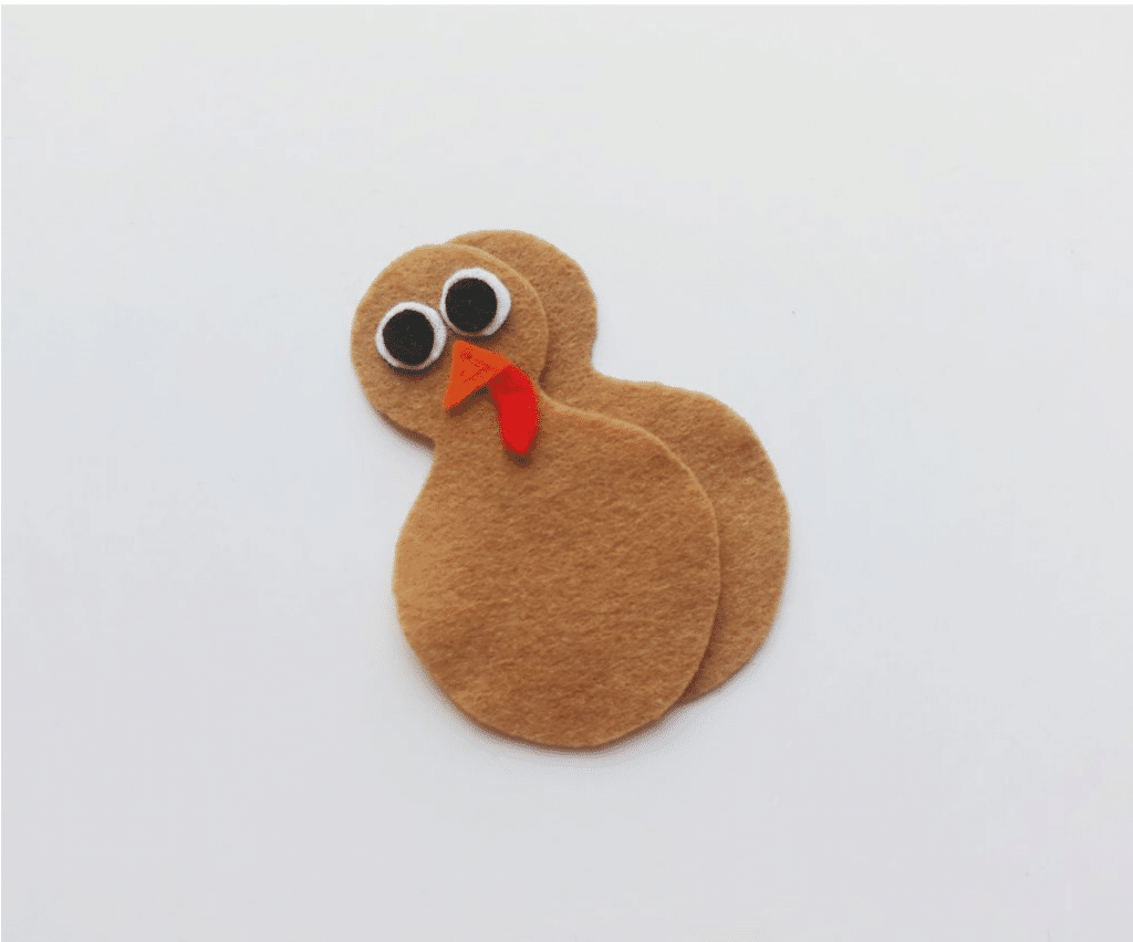 A felt turkey craft with two brown body parts, large black and white eyes, and a red felt wattle, placed on a white background.