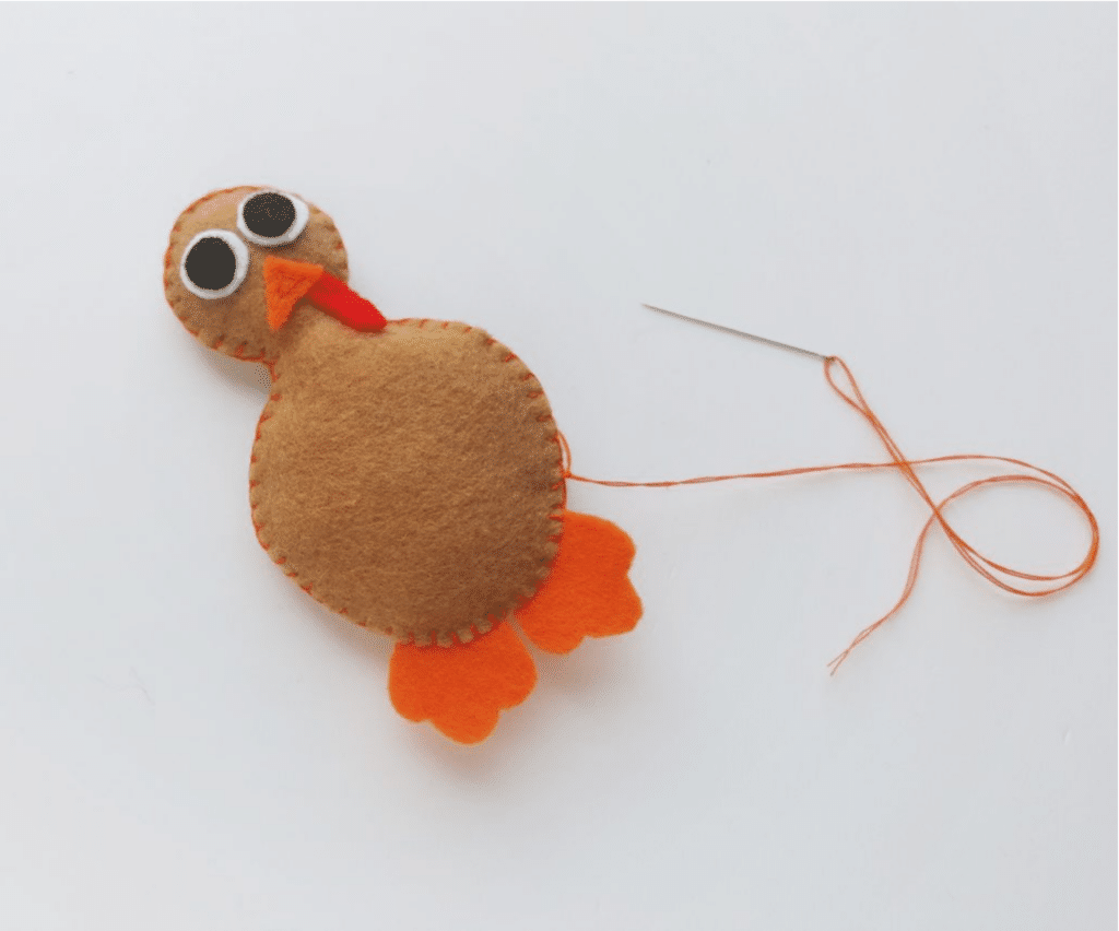 A turkey craft in progress, with orange and brown felt, black and white eyes, and an orange string attached to a needle for stitching.
