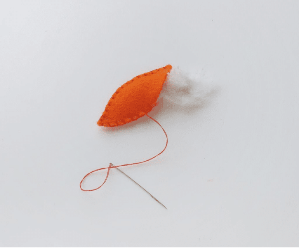 An orange piece of fabric with visible stitches and white stuffing, attached to a needle with orange thread, sits on a white background, resembling the beginnings of a charming turkey craft.