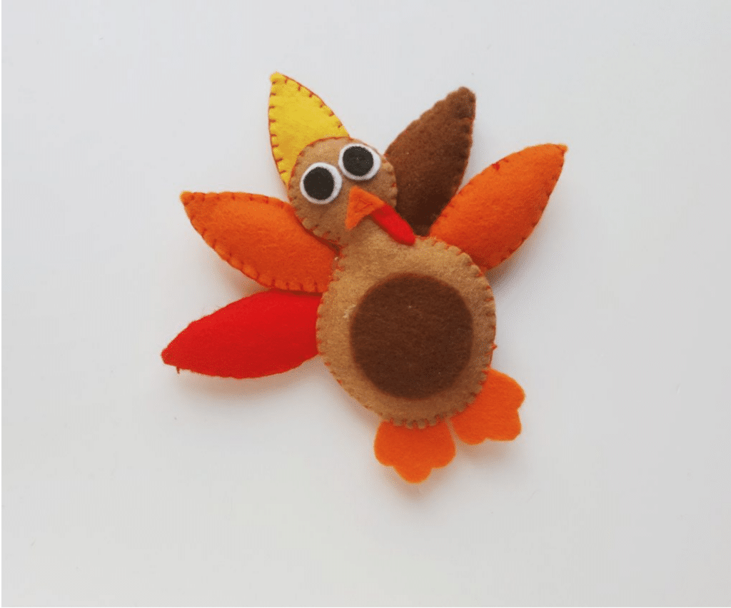 A turkey craft made from felt in the shape of a turkey with colorful feathers, featuring brown, orange, red, and yellow segments, and large round eyes on a white background.
