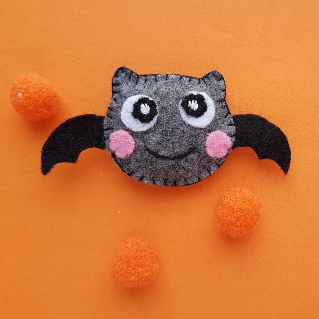 A grey felt bat with pink cheeks and black wings, surrounded by three orange pom-poms on an orange background.