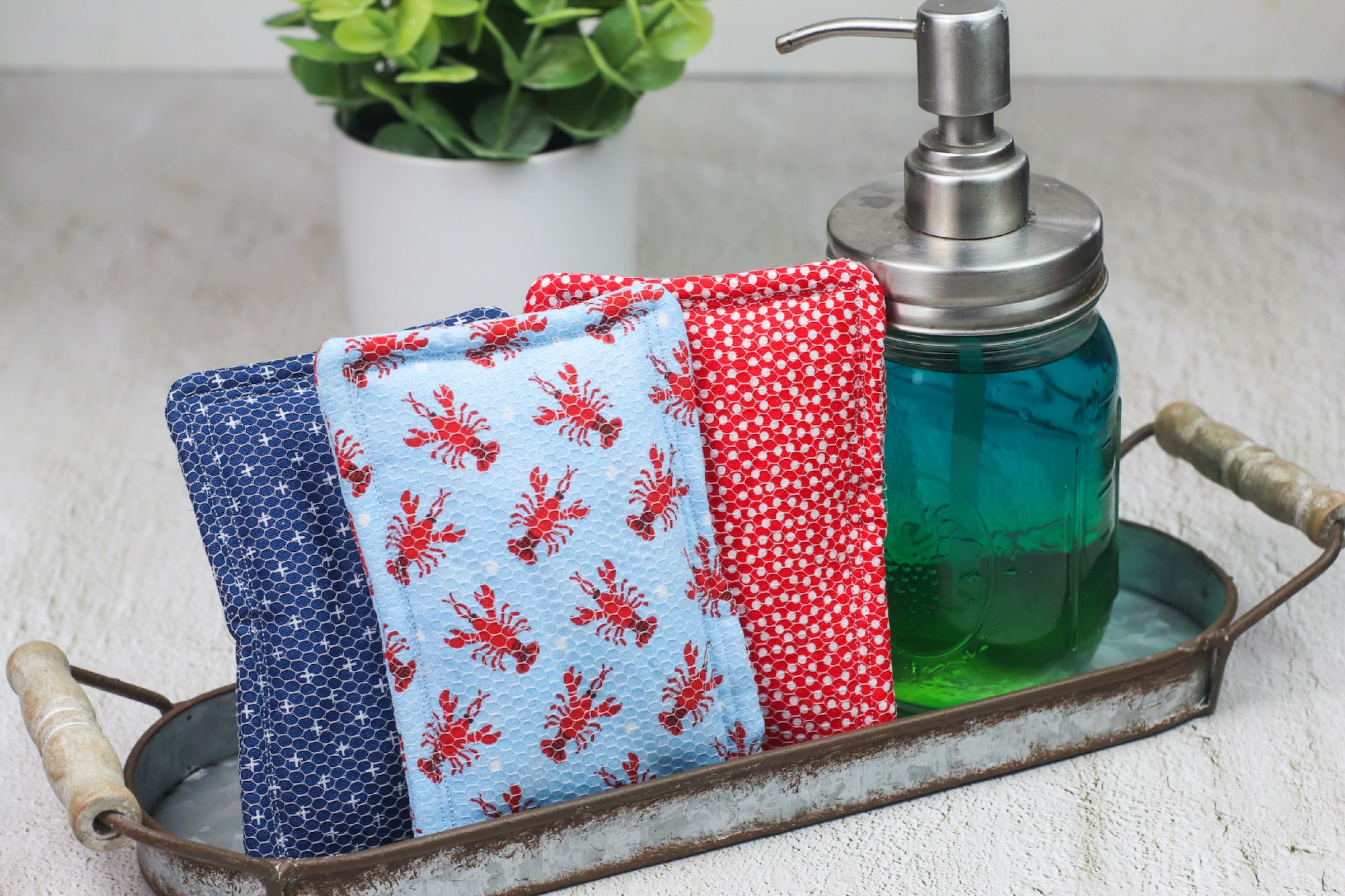 A metal tray holds a teal soap dispenser, a reusable sponge, and three folded towels with floral and polka dot patterns, set on a light surface.