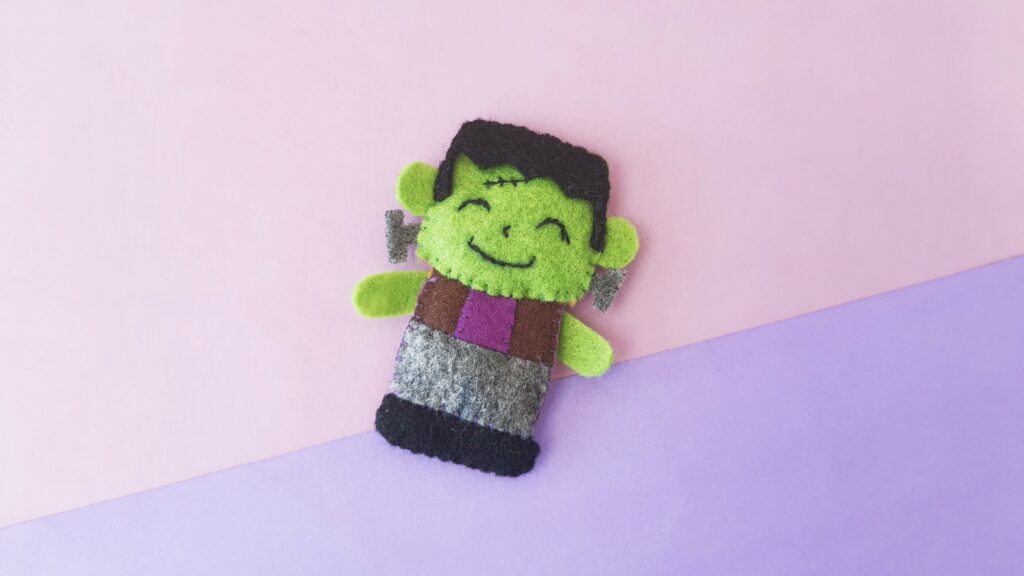 A felt frankenstein doll on a pink and purple background.