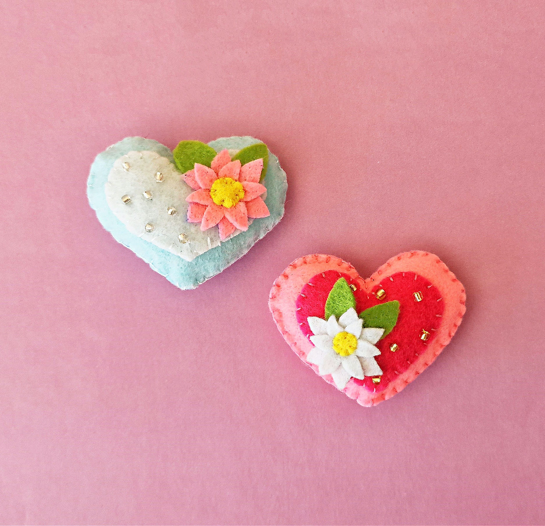 Two heart shaped felt brooches on a pink background.