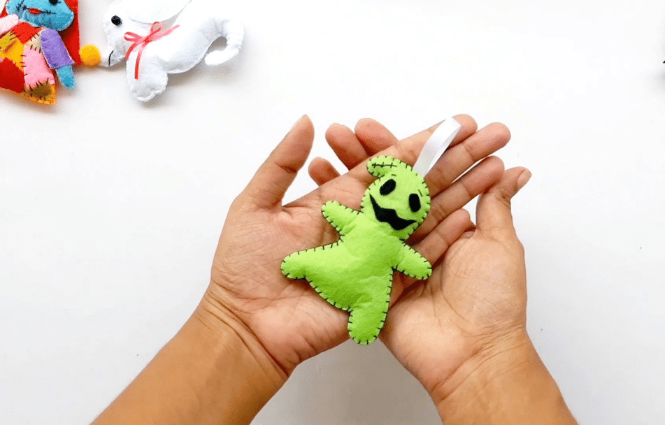 two hands holding a green stuffed felt oogie boogie against a white background 