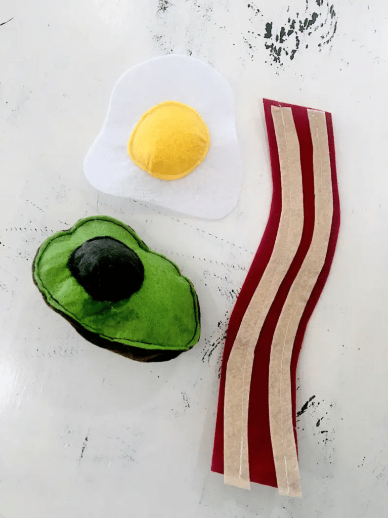 A piece of bacon, egg and avocado on a white table, crafted with attention to detail using felt food patterns.