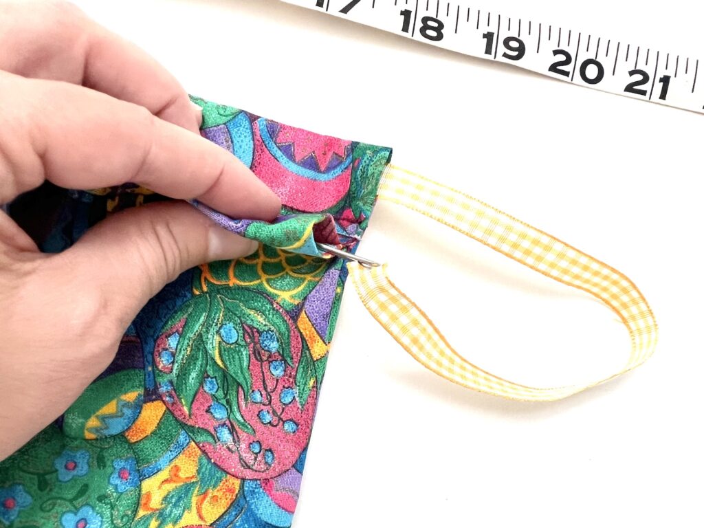 A hand sewing a colorful crayon bag with a patterned design, using a needle with a yellow ribbon attached, next to a ruler showing inches.