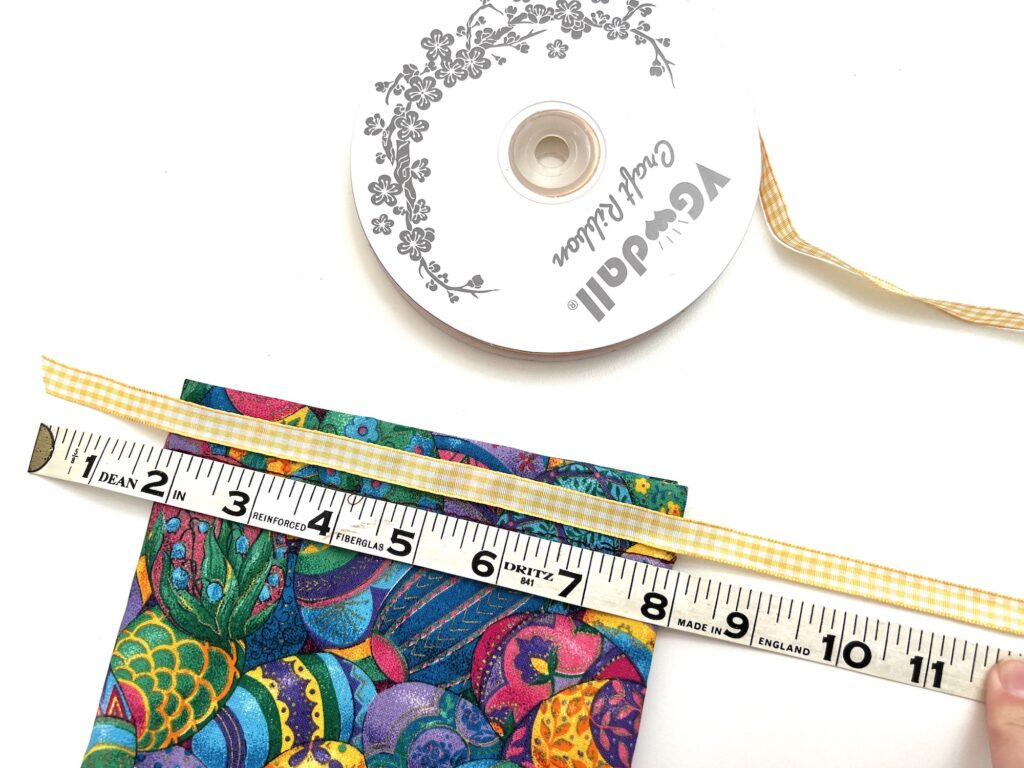 A hand holding a measuring tape next to a decorative ribbon and a crayon bag with floral patterns on a white surface.