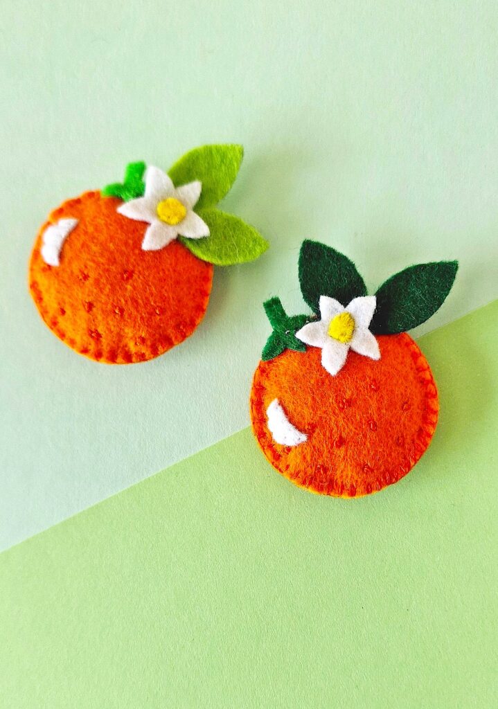 Two handmade felt orange crafts with white flowers and green leaves on a dual-toned background.