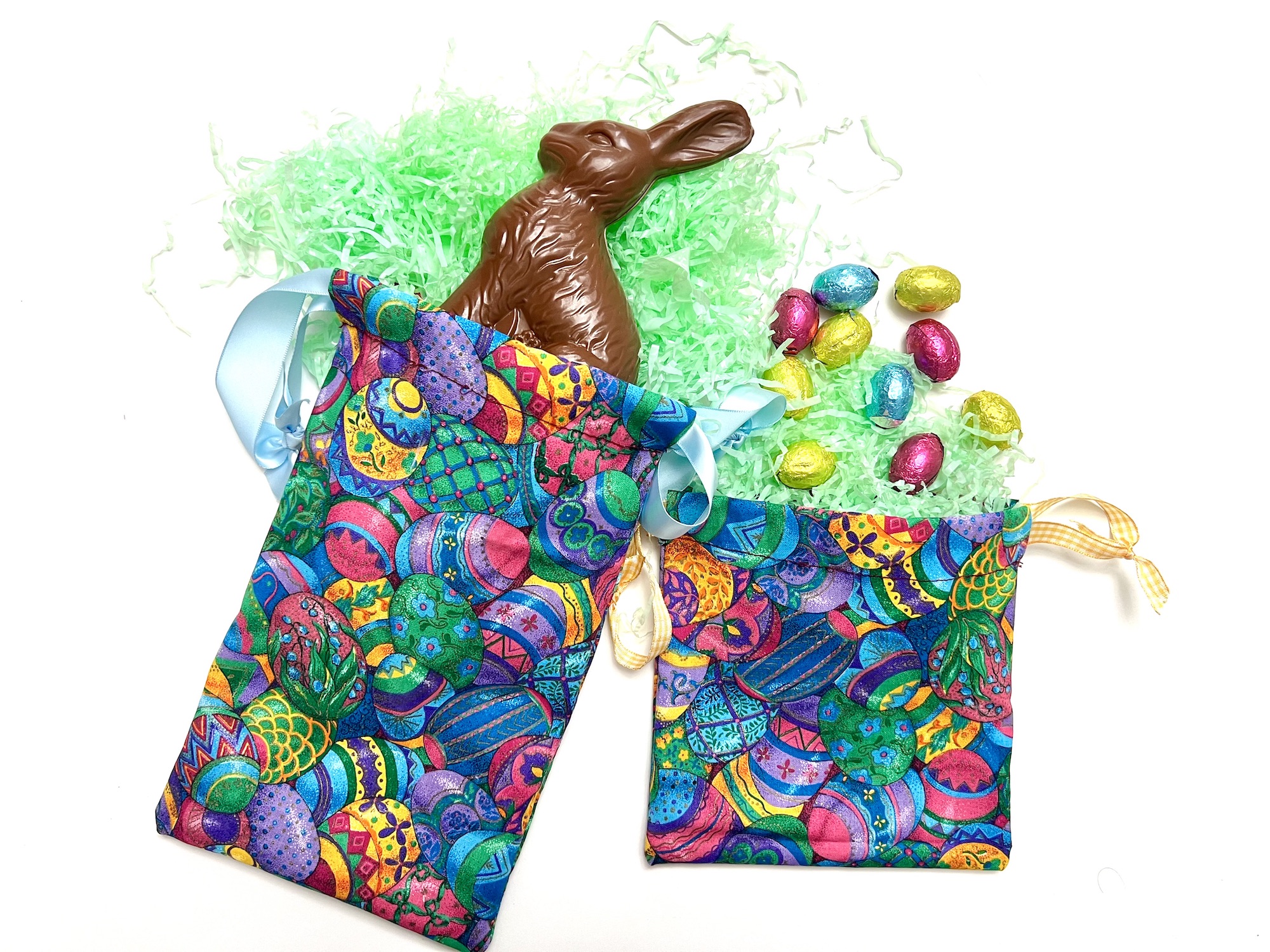 Two colorful Easter-themed crayon bags with patterned eggs, filled with green decorative shreds, assorted chocolate eggs, and a chocolate bunny.