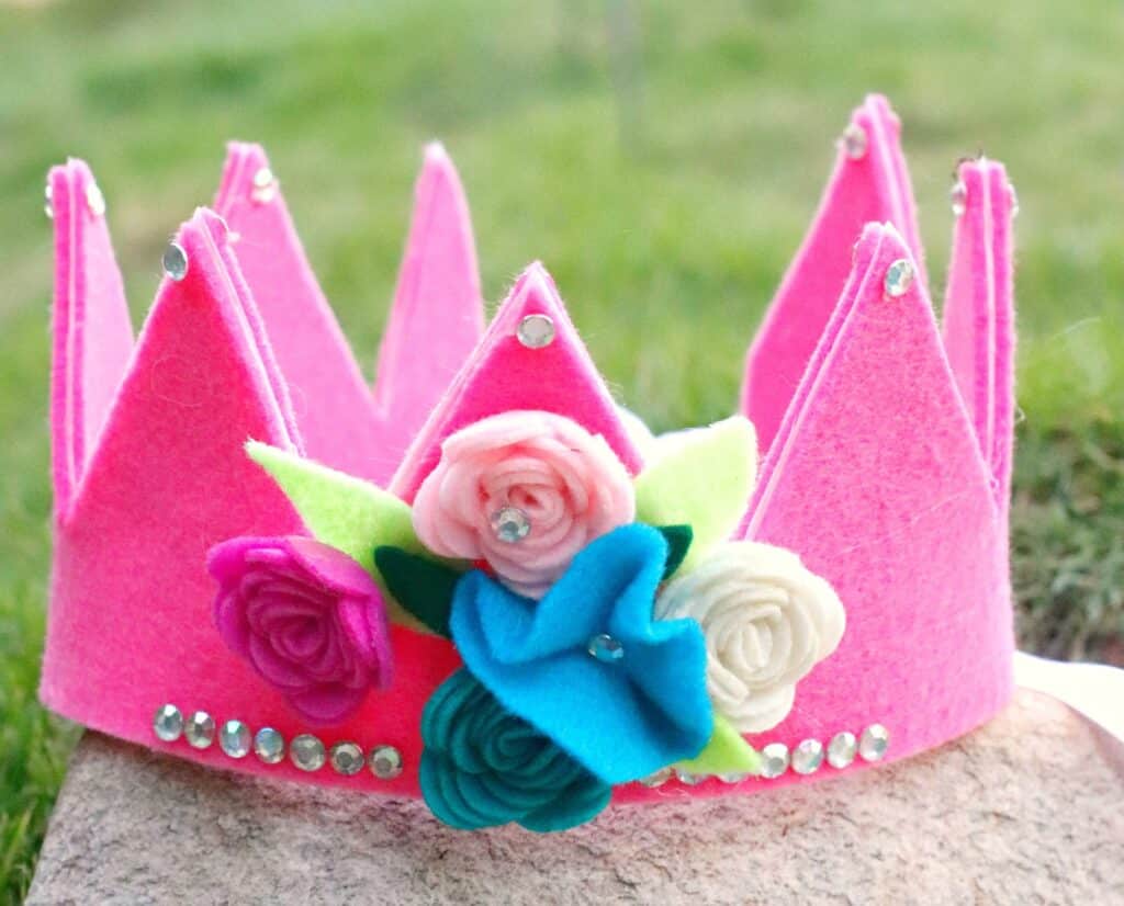 A pink felt crown decorated with rhinestones and fabric flowers rests on a stone, with grass in the background—perfect for an Operation Christmas Child gift box.