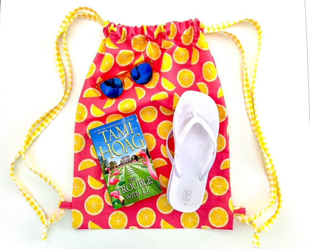Brightly colored drawstring backpack with citrus fruit pattern containing a pair of sunglasses, a book by Tami Hoag, and a single white flip-flop.