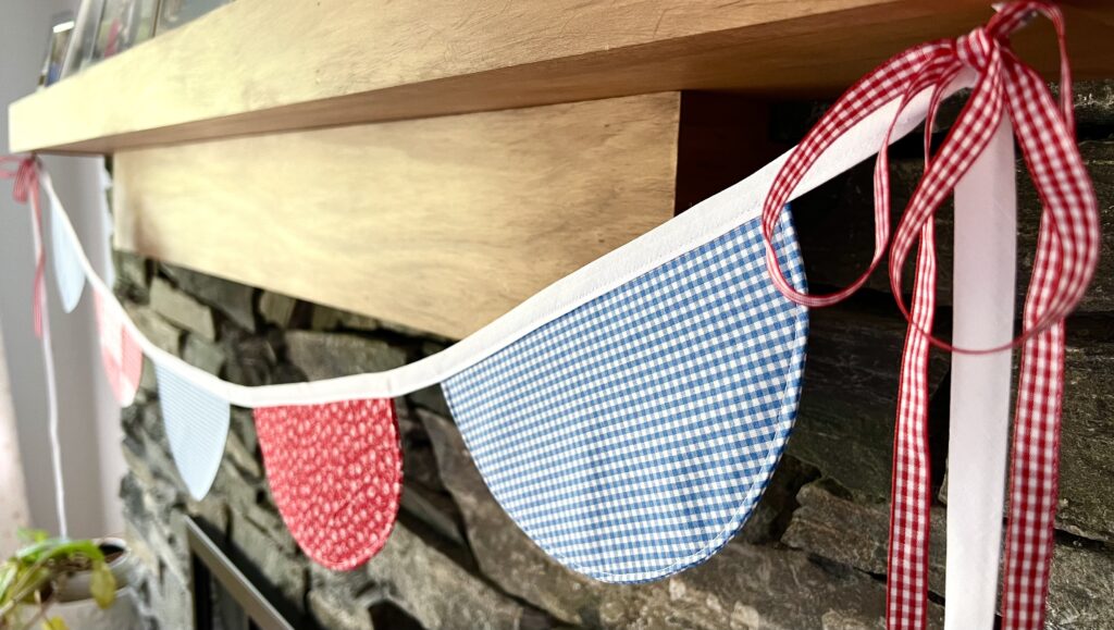 Decorative string of DIY blue and red gingham fabric bunting banner tied with checkered ribbons, hanging from a wooden beam against a stone wall background.