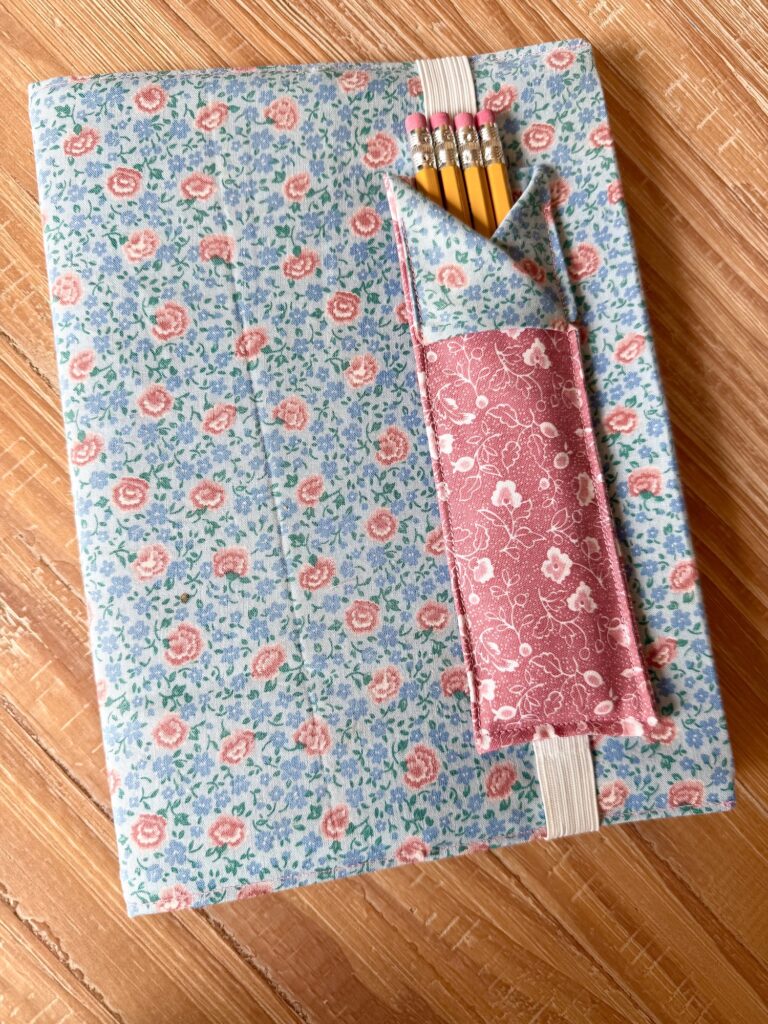 A fabric-covered notebook with a blue and pink floral pattern lies on a wooden surface. A pink pen holder bookmark on the cover holds several sharpened yellow pencils.