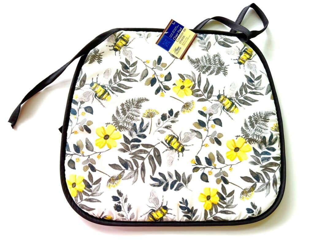 A square cushion with a bee and flower pattern on a white background. It has black trim and tie strings for attachment. A blue and yellow tag is attached to the top.