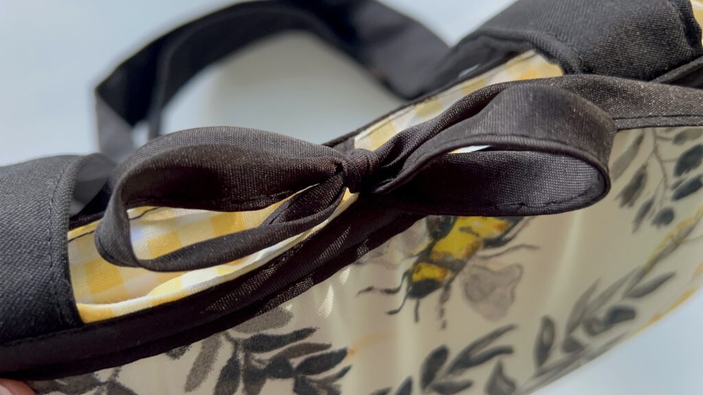 Close-up of a black bow on a handbag with a yellow and white checkered interior and floral-patterned exterior featuring a bee design.