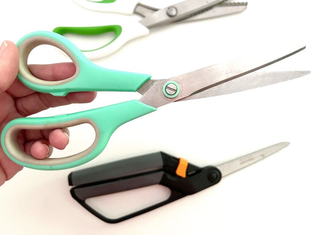 A hand holding green-handled sewing scissors, with another pair of white-handled scissors and a third pair of black-handled sewing scissors visible in the background.