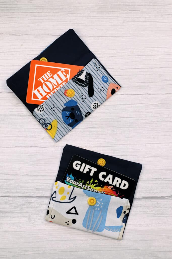 Two fabric wallets on a light wood surface, each open to reveal a gift card inside. The upper wallet holds a Home Depot gift card, perfect for DIY enthusiasts, while the lower wallet holds an Amazon gift card. These are thoughtful sewing gifts for men who appreciate both style and utility.