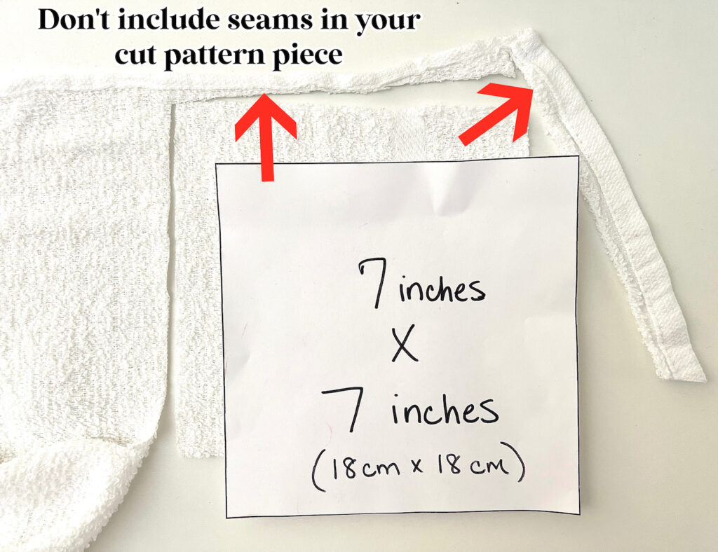 A piece of white fabric with a paper labeled "7 inches x 7 inches (18 cm x 18 cm)" sits ready for crafting reusable paper towels, alongside a note saying "Don't include seams in your cut pattern piece," accented with red arrows pointing at the seams.