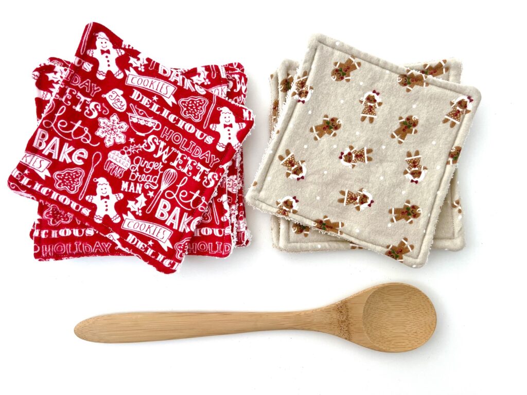 Two stacks of holiday-themed fabric coasters, adorned with gingerbread and cookie designs, sit beside a wooden spoon on a white background, perfectly complementing your kitchen's cozy atmosphere. Pair them with reusable paper towels for an eco-friendly festive touch.