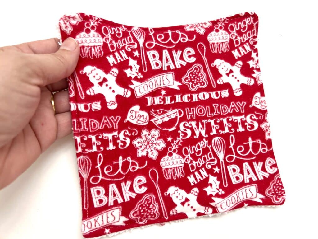 A hand holds a red fabric square adorned with white holiday-themed words and gingerbread designs, reminiscent of festive reusable paper towels.