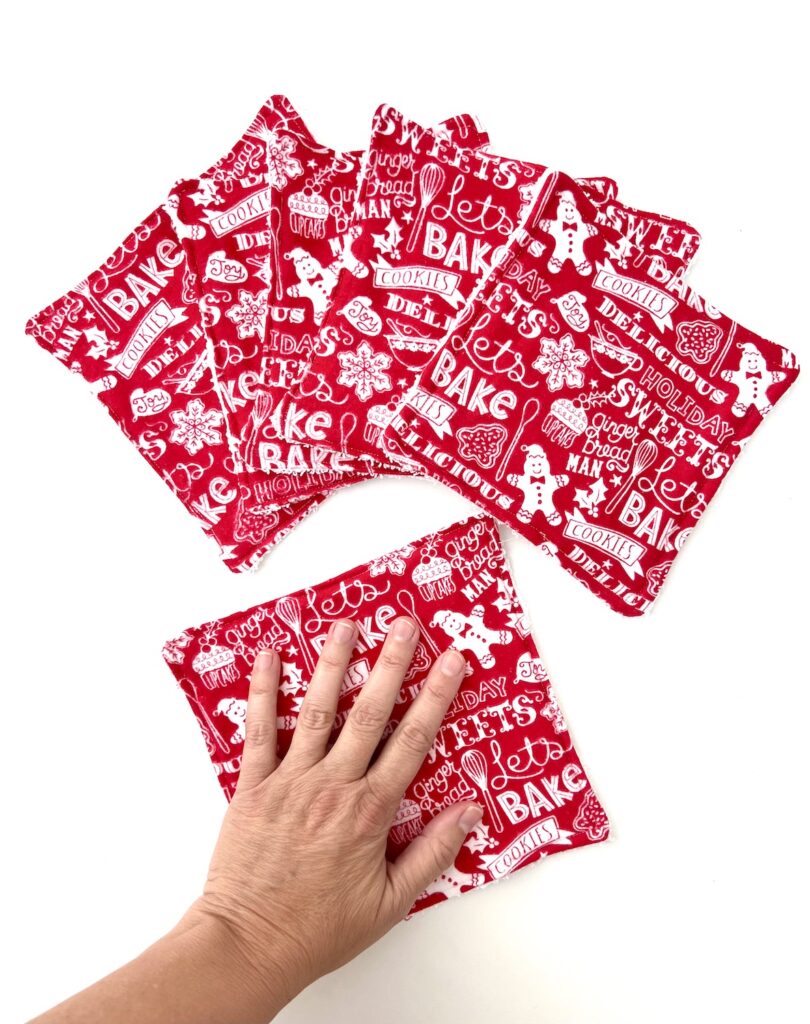 A hand holding a red potholder adorned with festive holiday-themed text and illustrations, accompanied by five matching potholders spread out on a white surface. Beneath them, the soft texture mimics the charm of reusable paper towels.