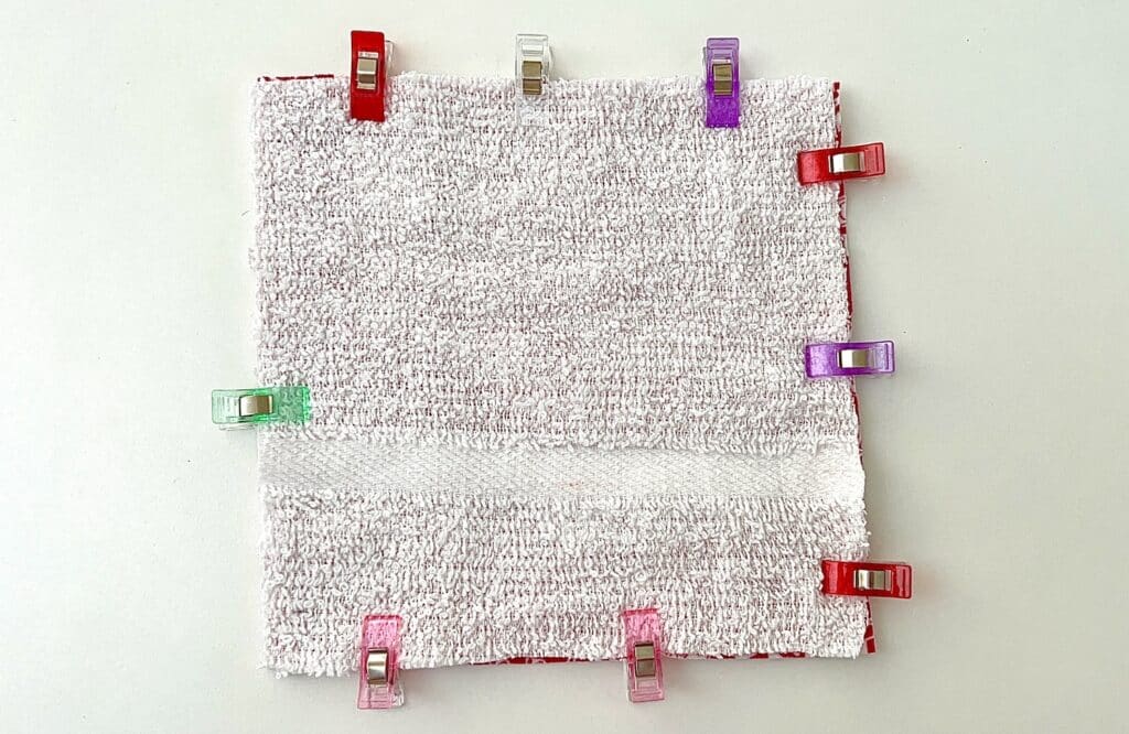 A square piece of white terry cloth, resembling reusable paper towels, is held by eight colorful clips on a flat surface.