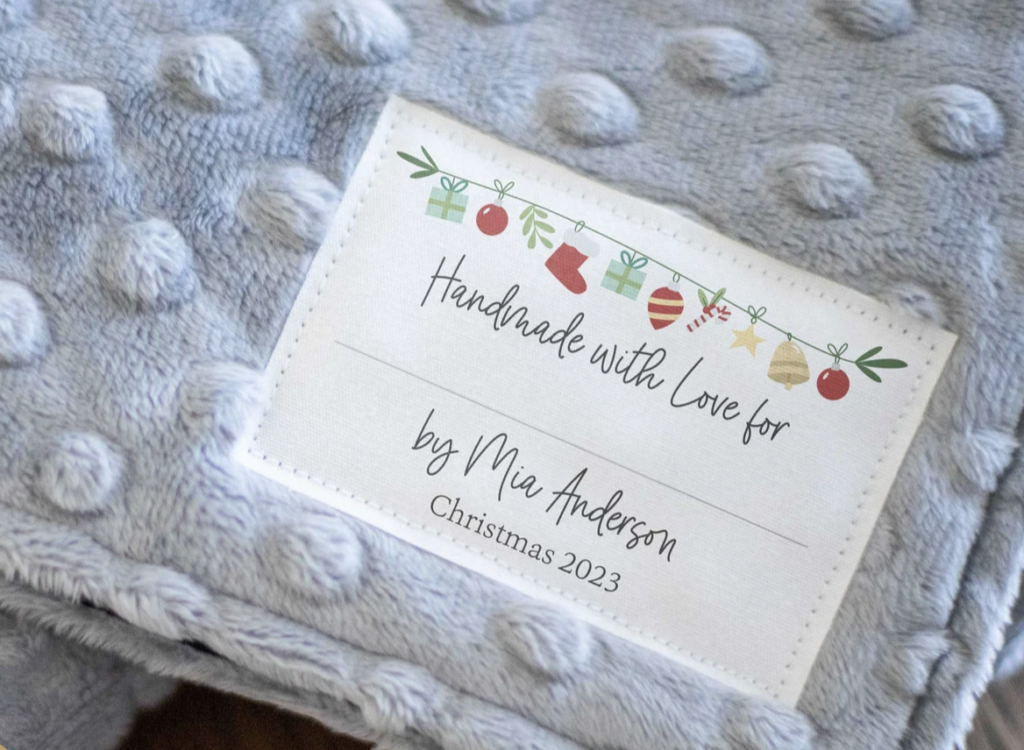 Close-up of a fabric label on a textured blanket. The label reads: "Handmade with Love for Mia Anderson, Christmas 2023." It features festive icons like presents and ornaments.