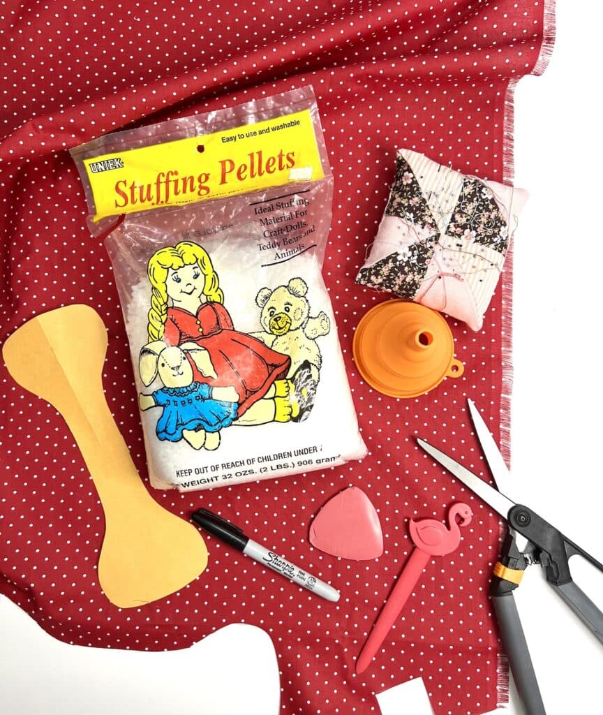 Craft materials including stuffing pellets, fabric, scissors, markers, and a pin cushion are arranged on a red polka-dot fabric.