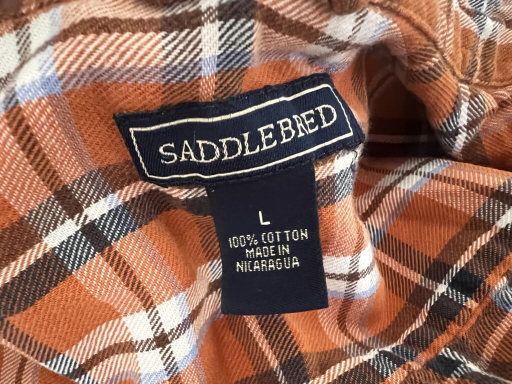 Close-up of a plaid fabric label reading "Saddlebred" and "100% cotton made in Nicaragua, L." The orange, brown, and white fabric is perfect for creating DIY hand warmers.