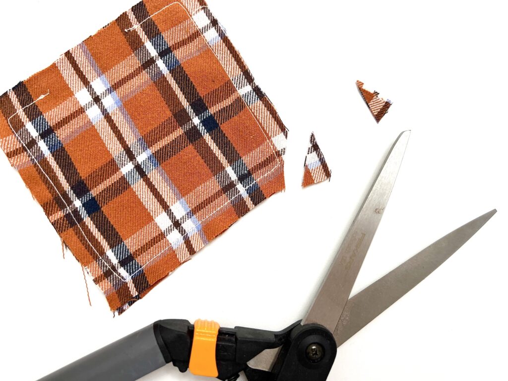 A pair of scissors rests beside a cut piece of orange and brown plaid fabric on a white surface, ready for crafting cozy DIY hand warmers.