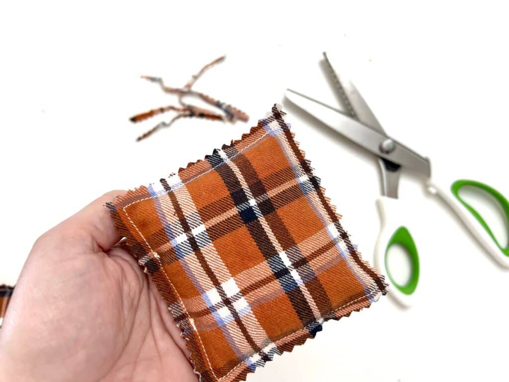 A hand holding a small plaid fabric pillow beside green-handled scissors on a white surface, perfect for crafting DIY hand warmers.