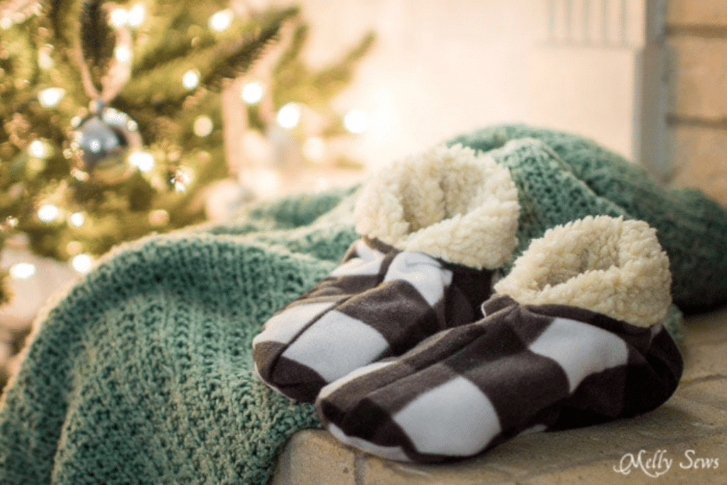 Cozy slippers with checkered pattern and fluffy lining placed on a green knit blanket, next to a lit Christmas tree.