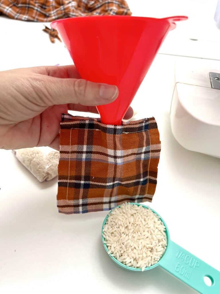 A hand craftily holds a red funnel over brown plaid fabric, ready for a DIY hand warmer project. In the foreground, a turquoise measuring cup filled with rice awaits its turn to bring comforting warmth.