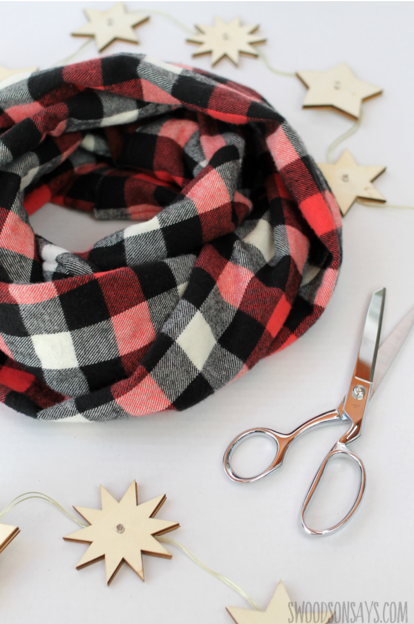 A black, red, and white plaid infinity scarf is surrounded by wooden star ornaments and a pair of scissors on a white surface.