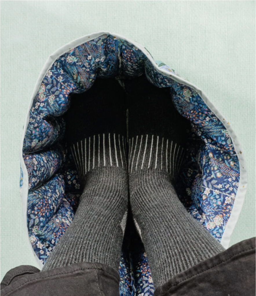 Feet wearing black and gray socks are cozily tucked into a round, quilted foot warmer with a blue floral pattern, making for one of the coziest sewing projects.