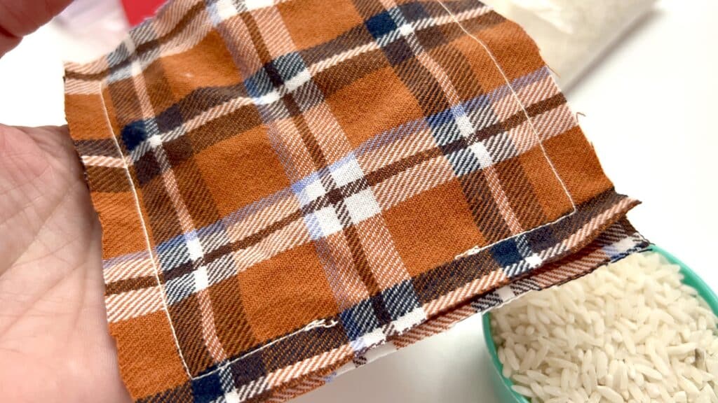 A close-up captures a person's hand holding a piece of orange, blue, and white plaid fabric near a bowl of uncooked rice—perfect for crafting personalized DIY hand warmers.