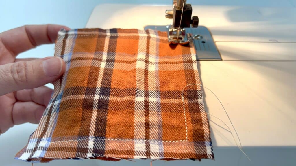 A hand guides a piece of brown plaid fabric under the needle of a sewing machine, crafting cozy DIY hand warmers for chilly days.