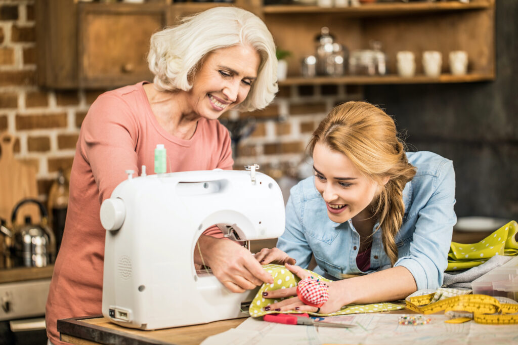 Two women, one older with short white hair and the other younger with long blonde hair, smile as they work on a sewing project at a wooden table.