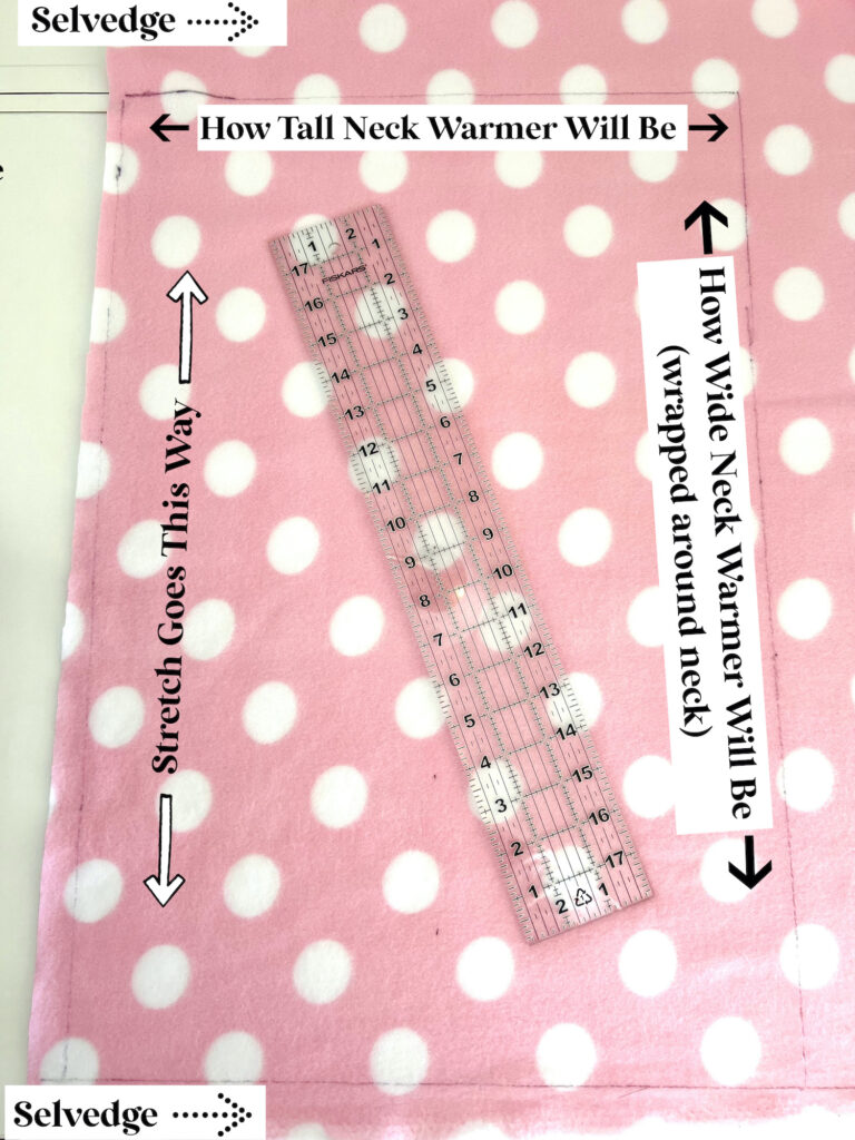 Pink fabric with white polka dots, a clear ruler on top, and labeled arrows indicating stretch direction, and dimensions for a neck warmer.