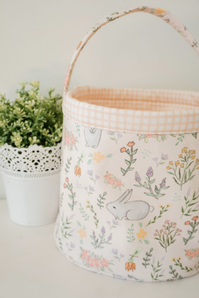 Fabric basket featuring charming bunny sewing patterns and a floral print, sitting beside a white pot filled with lush green foliage.