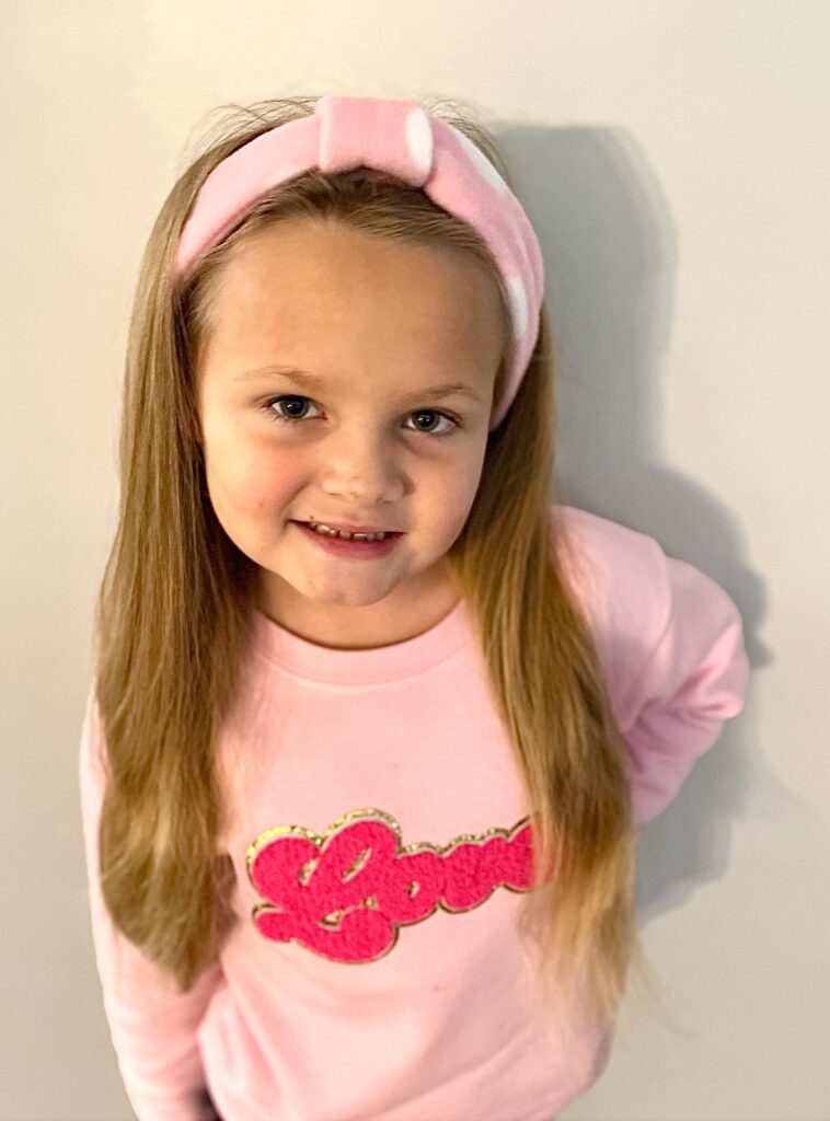 Young girl with long hair, wearing a pink fleece headband and matching sweatshirt with "Love" written on it, smiles standing against a plain backdrop.