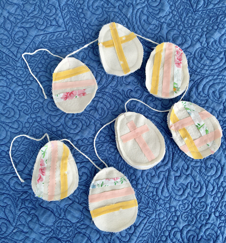 Fabric decorations resembling eggs with pastel stripes and cross patterns are artfully arranged on a quilted blue background, featuring whimsical bunny sewing patterns.