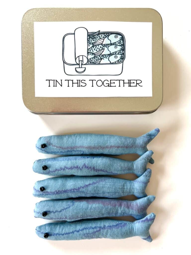 A tin adorned with a charming sardine sketch and the words "Tin This Together" below showcases five playful fabric fish toys neatly aligned in a row.