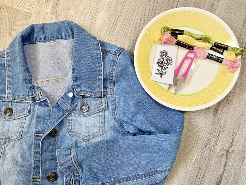 A denim jacket is on a wooden floor next to a plate with embroidery threads, scissors, and a floral pattern.