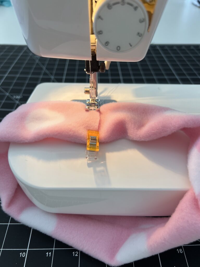 A sewing machine stitches pink fabric with a yellow clip on a black grid mat, crafting a cozy fleece headband.