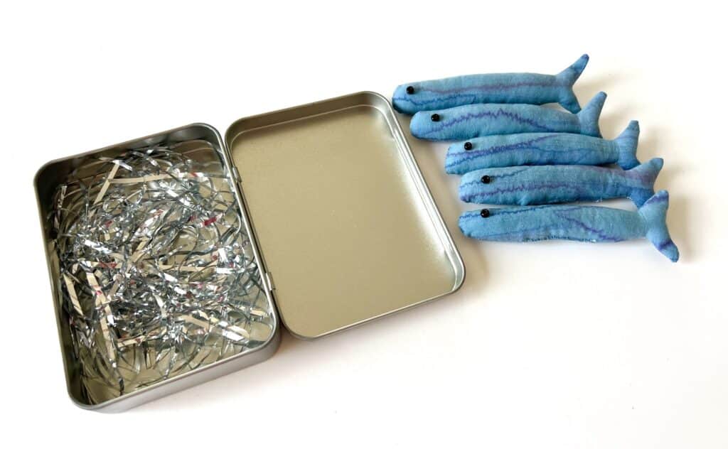 An open metal tin with shredded material inside evokes the essence of sardines, set next to five plush blue fish on a crisp white background.