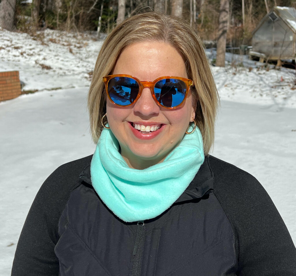 Person wearing sunglasses and a vibrant turquoise neck warmer smiles while standing on snow-covered ground.