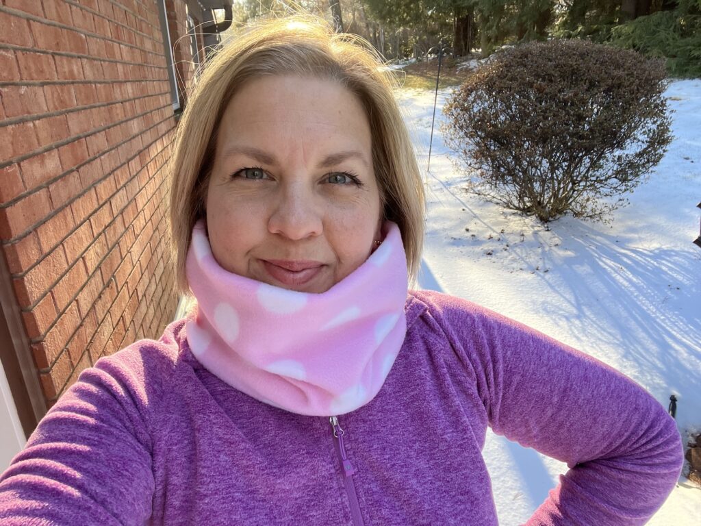 A person in a pink fleece jacket and polka dot scarf, with a cozy neck warmer, stands outside on a snowy day near a brick wall and shrubs.